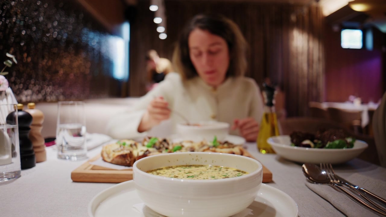 Donna che mangia zuppa a un tavolo di ristorante con una ciotola di zuppa cremosa in primo piano e pizza e insalata sullo sfondo.