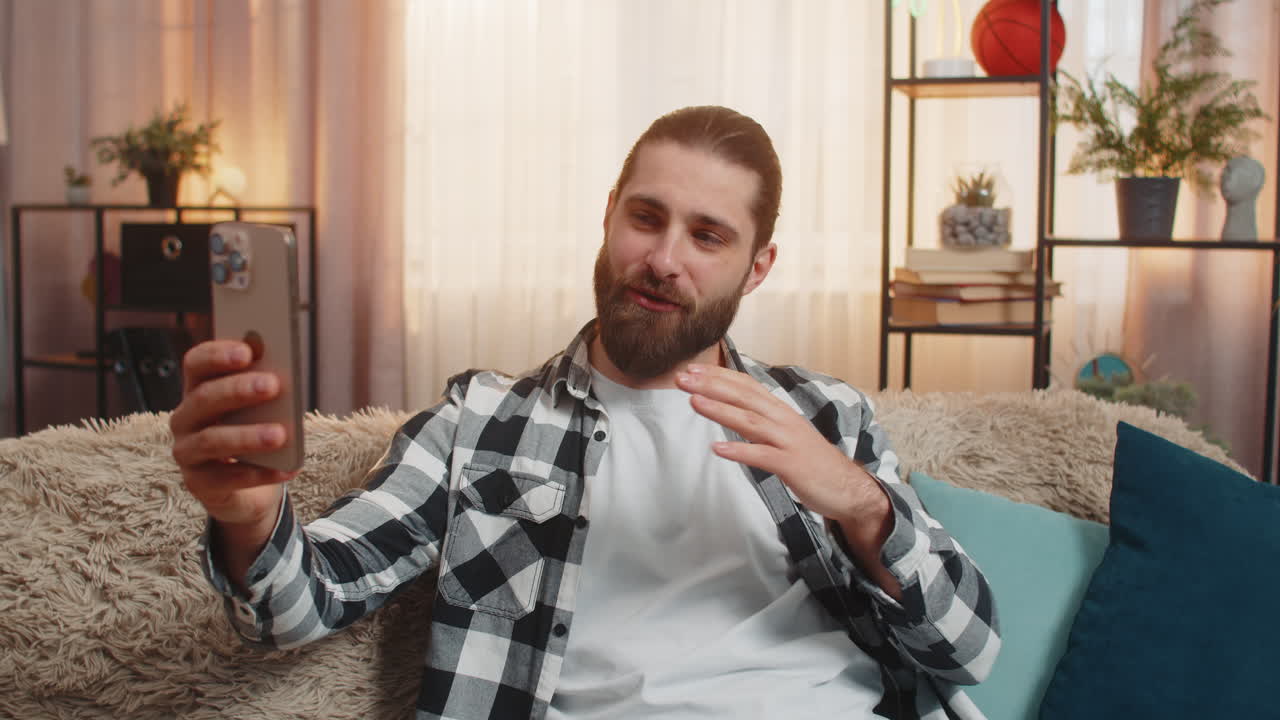Young man blogger at home sofa selfie mode waving to camera fun story casual influencer vibe