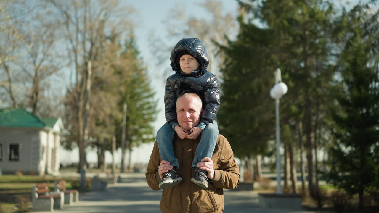 Father carrying his son on his shoulders both smiling, the father is wearing a brown jacket and beanie, and the child is in a black jacket with trees and light poles behind