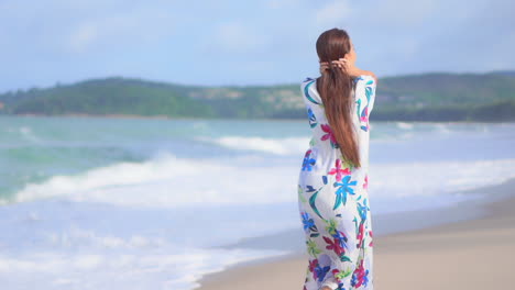 Back-to-the-Camera-While-walking-along-the-beach,-a-young-woman-pushes-back-her-windswept-hair