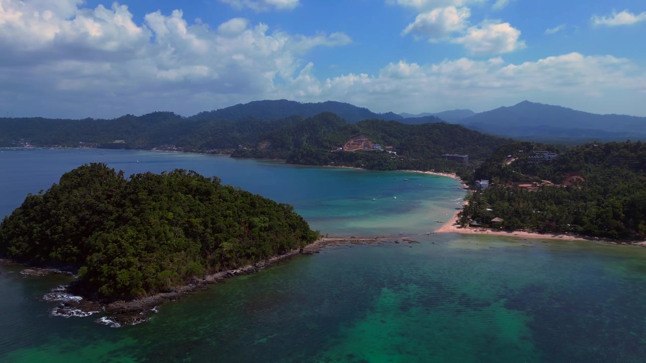 Depeldet Island's lush green landmass connecting to a white sand beach and coastal development, surrounded by clear blue waters. Great aerial view panorama drone overview