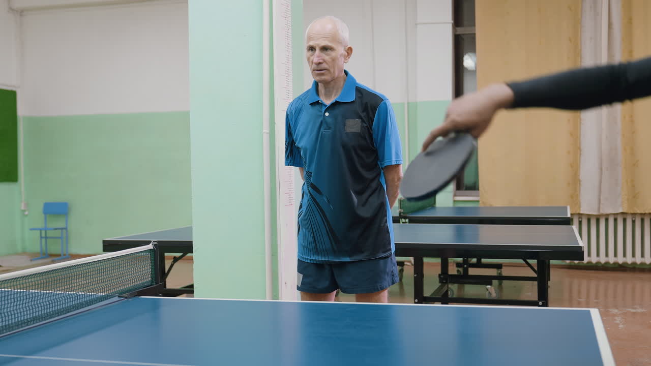 Senior coach in blue shirt attentively observes young player holding paddle at table tennis table, focusing on technique and preparation during indoor sports training session