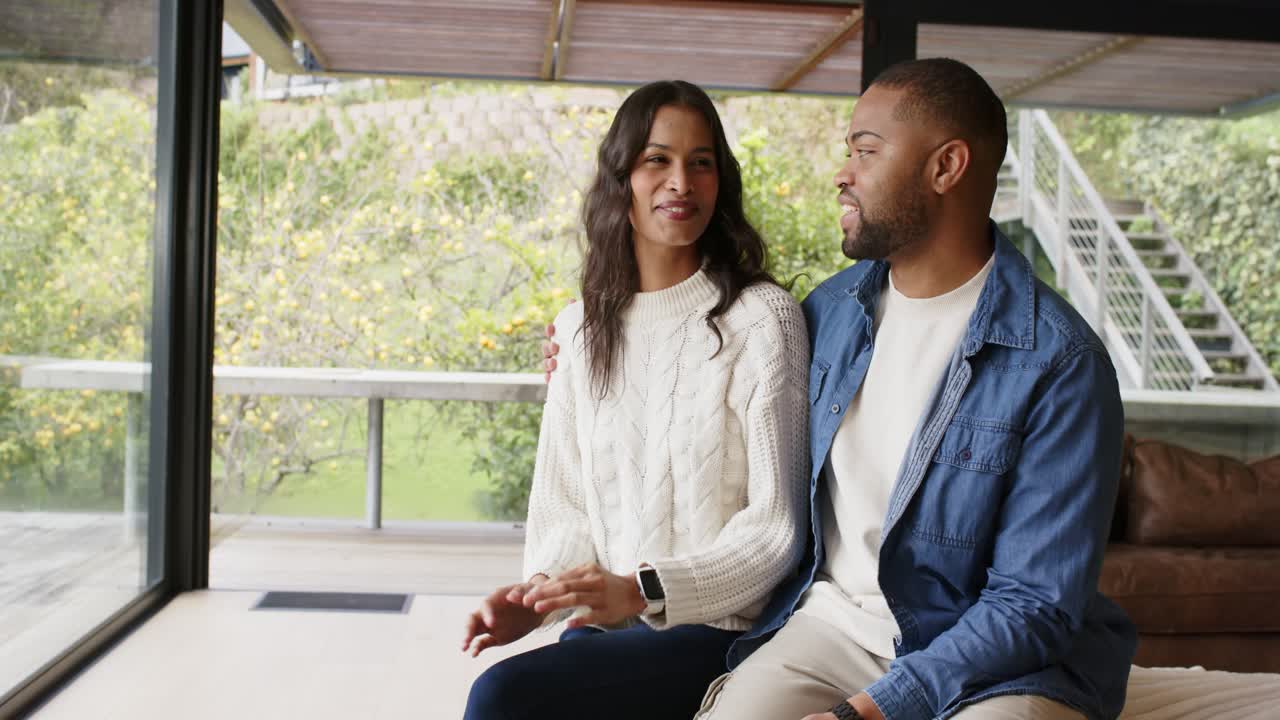 Diverse couple exchanging smiles and chatting on sofa before glass wall overlooking garden