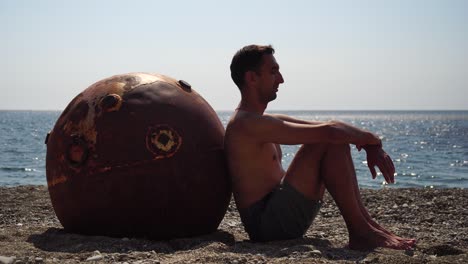 man doing yoga exercise outdoors near to an old rusty floating marine mine on the beach with rocky shore and sea background. healthy lifestyle, pollution, nature protection, war and peace concept