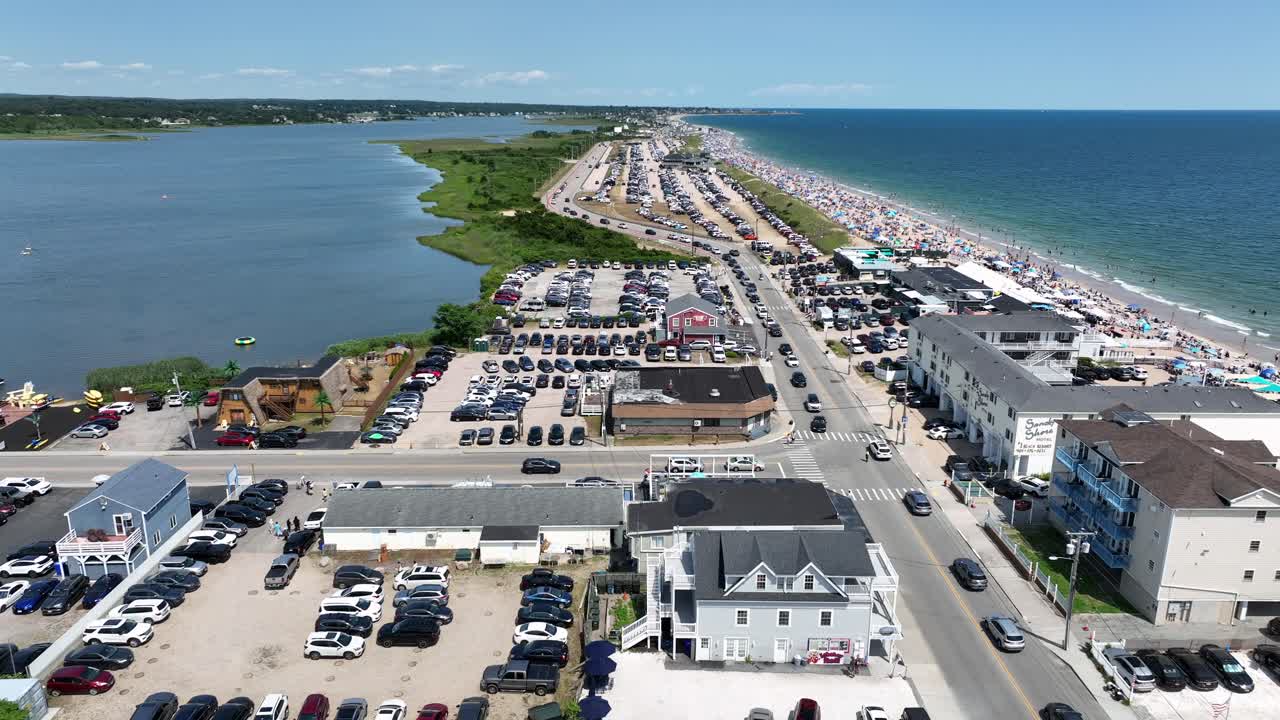Aerial view of Misquamicut Beach, Rhode Island. Crowded summer shoreline, parked cars, beachfront hotels, shops and the calm waters of Winnapaug Pond under bright blue sky