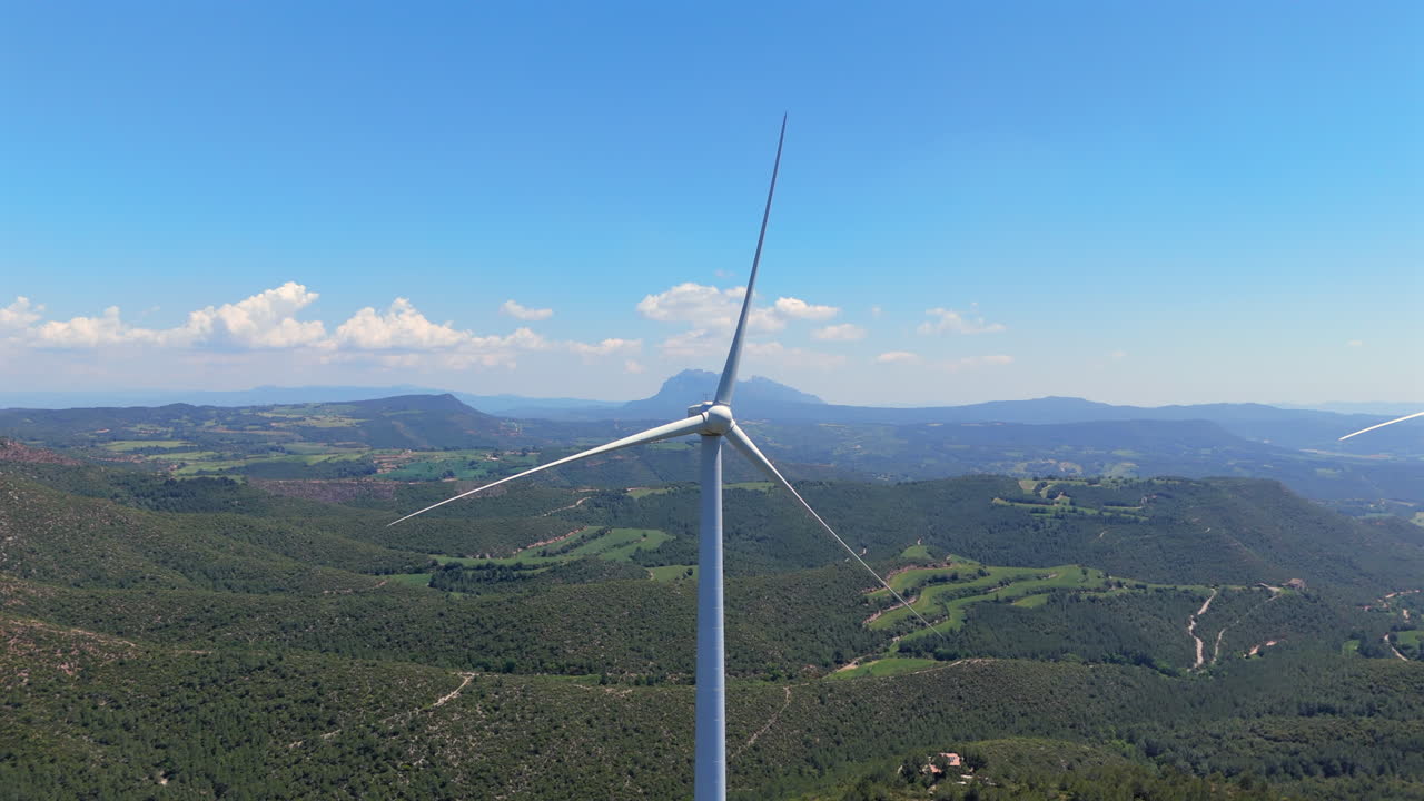 Drone flies straight toward a wind turbine over lush hills, ending very close to the spinning blades. Mountainous landscape, blue sky and green valleys emphasize renewable energy in nature