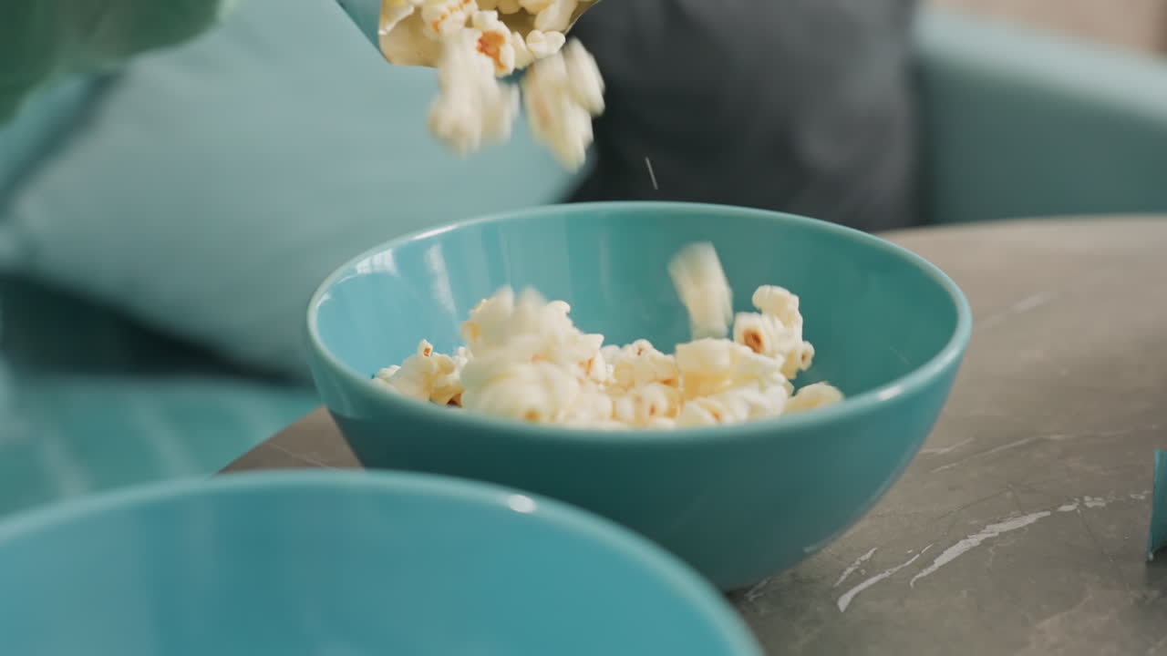 Primo piano di popcorn che cadono in una ciotola sul tavolo, ciotola blu sul tavolo, preparazione informale di uno spuntino, mani femminili che versano popcorn, ambiente cucina o soggiorno, momento spuntino, spuntino leggero.