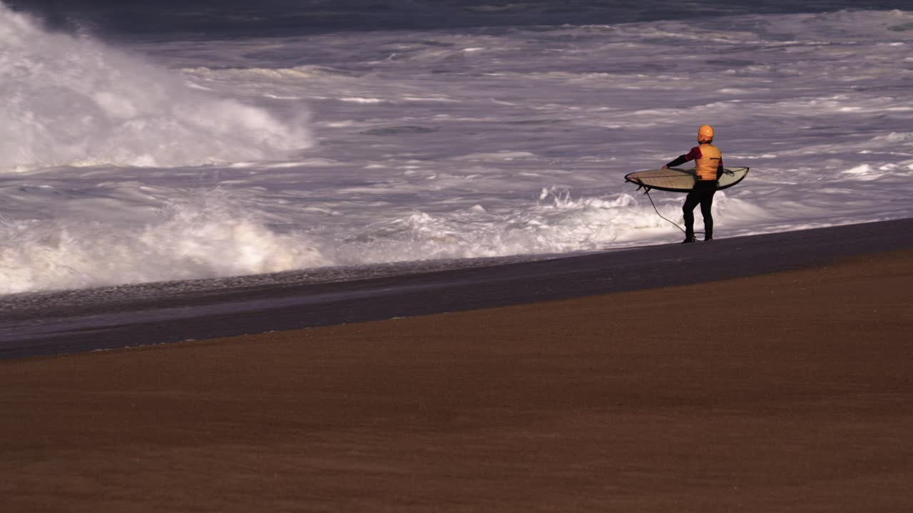 surfista con chaleco amarillo y casco con tabla, listo para ir al agua, parado en la orilla frente a olas salvajes