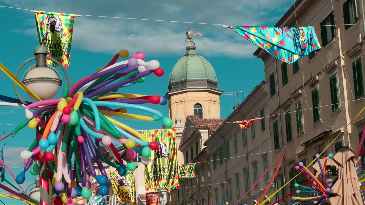 Slow motion nærbillede af farverige balloner, der hænger over en historisk gade i Rijeka. Gamle byarkitektur i baggrunden skaber en festlig karnevalsstemning på en solrig dag.
