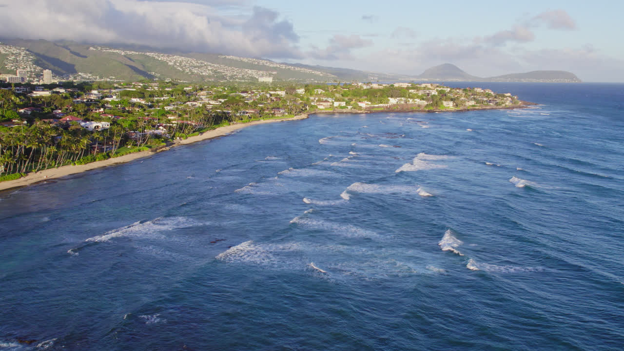 Onde con creste bianche sormontano le acque blu dell'oceano Pacifico lungo la costa dell'isola di Oahu, Hawaii, con villaggi costieri e vegetazione tropicale