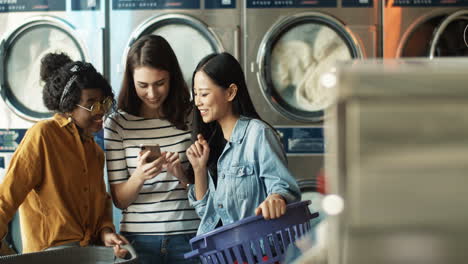 Girl Showing Photos On Smartphone To Mixed Races Female Friends While Washing Machines Working And Cleaning Clothes