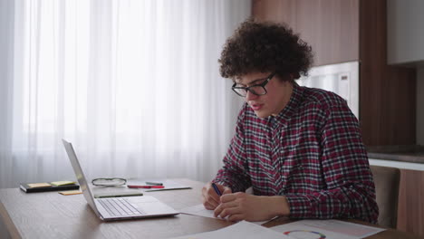 Curly-haired-Male-student-attractive-young-boy-in-glasses-is-studying-at-home-using-laptop-typing-writing-in-notebook.-College-student-using-laptop-computer-watching-distance-online-learning-seminar