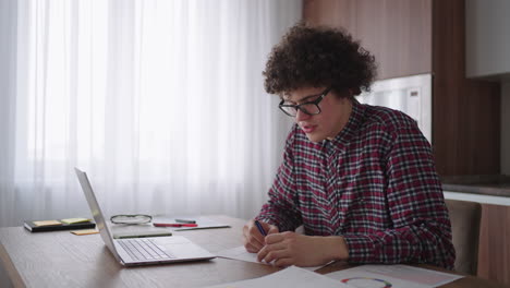 curly haired male student attractive young boy in glasses is studying at home using laptop typing writing in notebook. college student using laptop computer watching distance online learning seminar