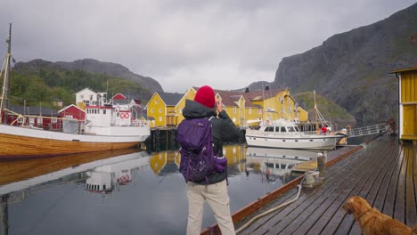 fotografía en cámara lenta de una joven turista fotógrafa y su perro golden retriever fotografiando el hermoso pueblo de pescadores de å y sus casas de rorbu rojo en las islas lofoten, noruega