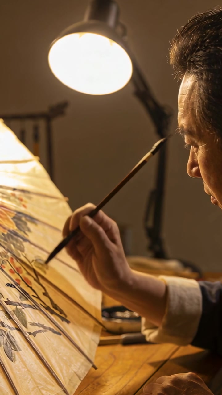 Vertical video: Lamp lighting man decorating parasol at bench with brush, rolled cuffs, copy space
