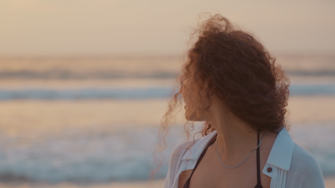 Ritratto di donna contemplativa sulla spiaggia di un'isola al tramonto, capelli ricci che catturano la luce dorata, onde delicate che lambiscono la riva, silhouette di un'isola lontana, brezza leggera, umore riflessivo, lenta panoramica cinematografica