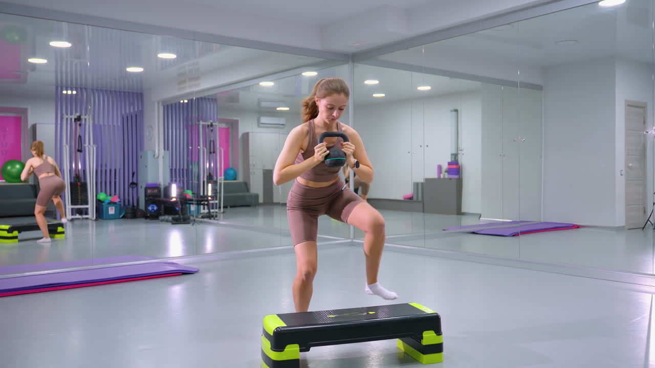 여성은 무게를 들어 올리고, 에어로빅 벤치에 발을 고, 거울 벽이 있는 체육관에서 다시 무게를 들고, 근력 훈련과 심장 운동 틴을 결합하여 근육을 구축합니다.