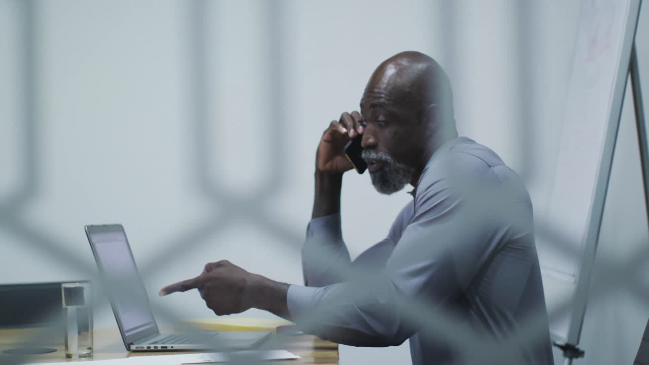 African american businessman talking on smartphone, using laptop sitting in office conference room
