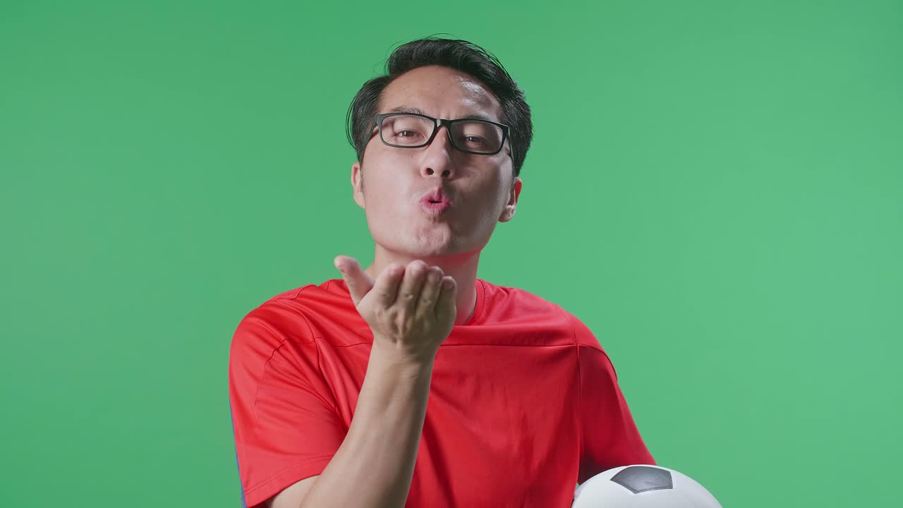 Close Up Of Asian Man With A Ball Blowing Kisses And Smiling To Camera While Cheering Soccer On Green Screen Background