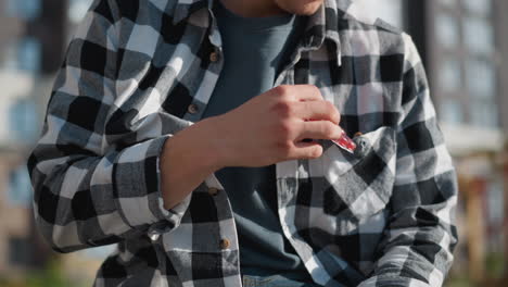 close up of man wearing checkered shirt reaching into front pocket to retrieve drug tablet during sunny day outside with blurred view of urban structures and warm natural light on skin and fabric