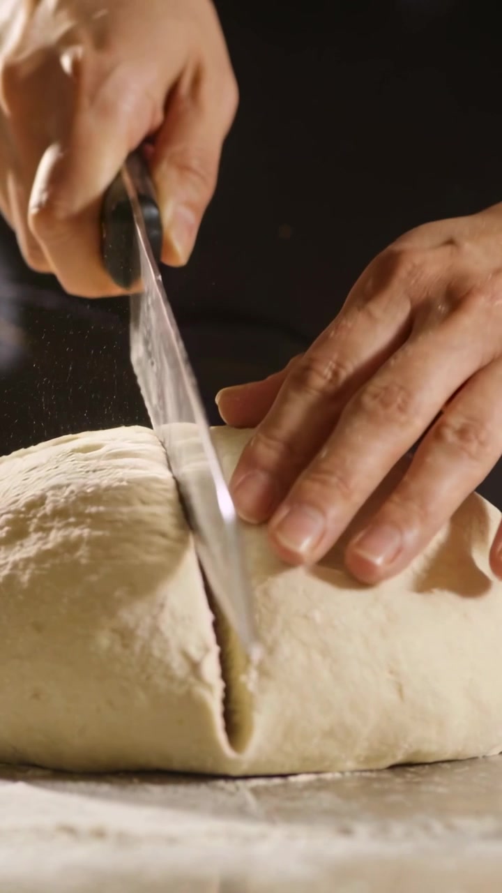 Vertical video: Placing knife, hands scoring proofed loaf on floured board, guiding cut for baking
