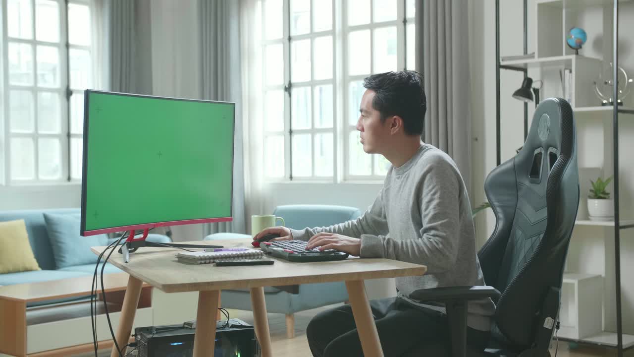 Asian Man In Long Sleeved T-Shirt And Black Pants Typing On Green Screen Desktop Computer While Working At Home.