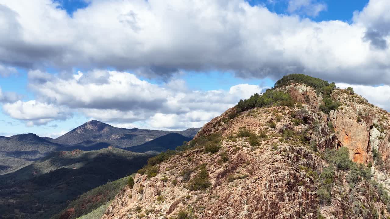 Camera smoothly pans along rugged Split Rock ridge, revealing expansive mountain landscape under dynamic clouds and bright daylight in Warrumbungle National Park, Australia