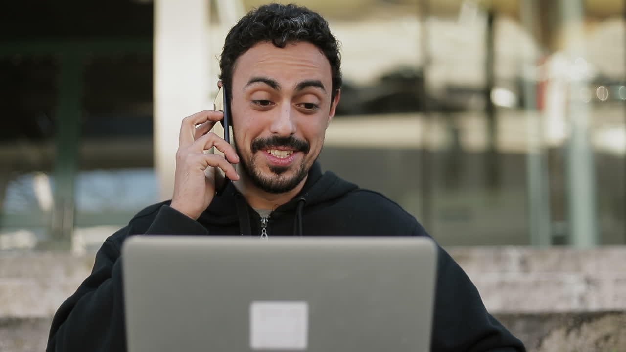 vista frontal de un hombre árabe con cabello rizado oscuro y barba con capucha negra sentado en las escaleras afuera