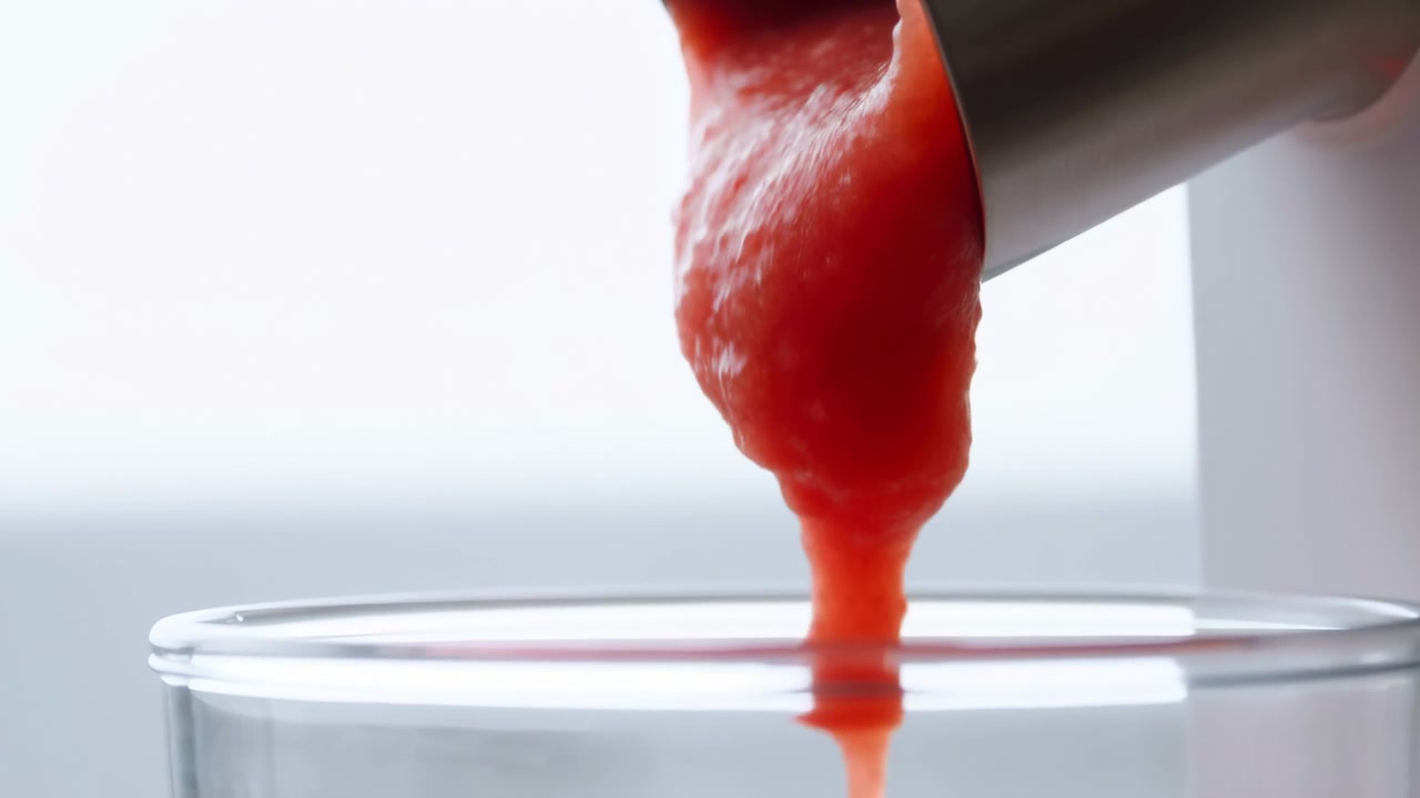 Scena in movimento che mostra una densa purea rossa che scorre da un beccuccio corto in acciaio inossidabile in un bicchiere trasparente, con un flusso lucido e strutturato, una goccia arrotondata che si forma vicino al bordo, riflessi tenui e un aspetto da studio pulito