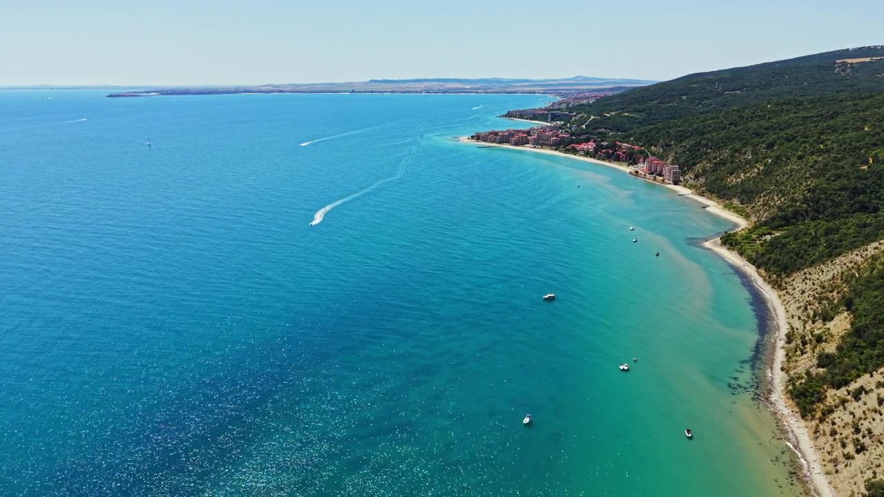 Vista di una costa con acqua blu cristallina, colline verdi e barche sull'acqua durante una giornata di sole splendente vicino a una città costiera.