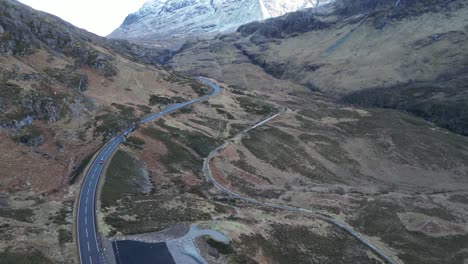 Camino-Sinuoso-En-Glencoe-Con-Montañas-Nevadas-En-La-Distancia,-Clima-Nublado,-Vista-Aérea