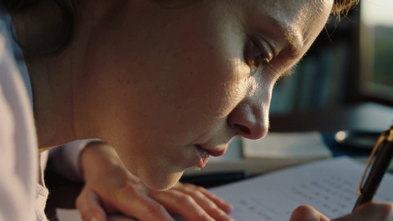 Writing woman in light blouse leaning over desk, pen touching paper and completing printed form