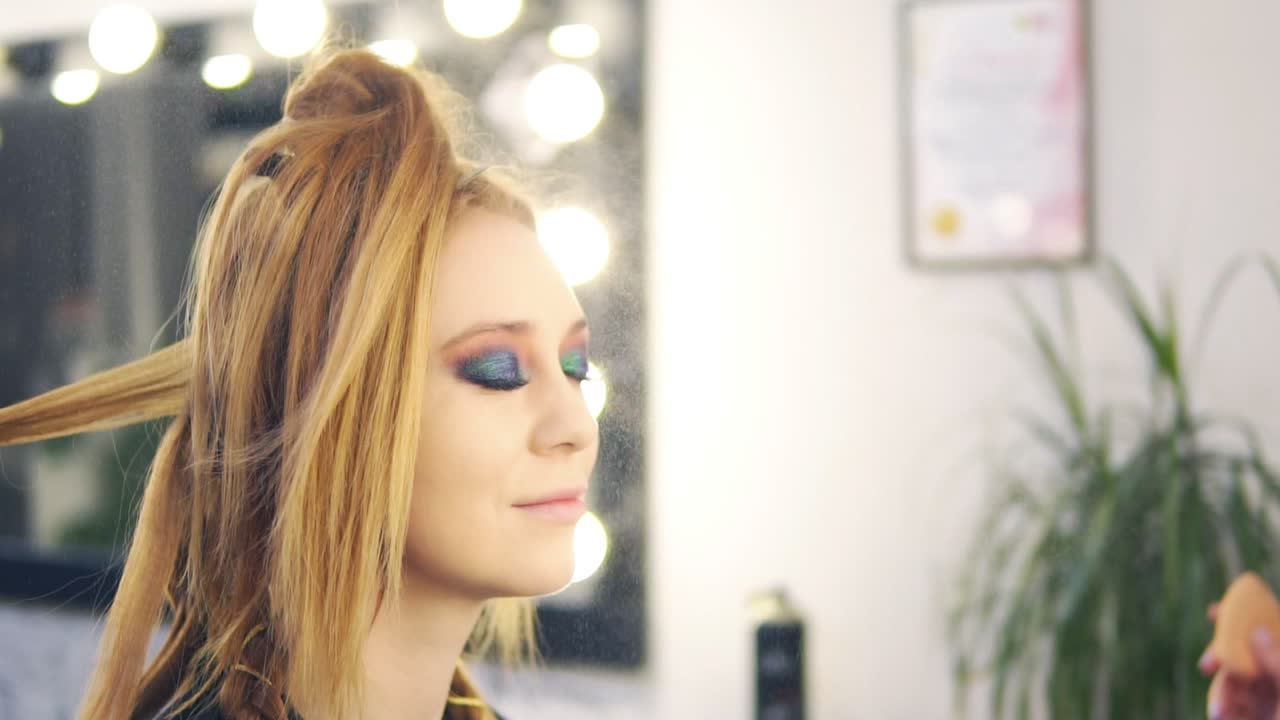 A blonde is sitting on the chair and a make up artist is fihishing her visage with a face spray. Her eyes are already finished. Hairdresser styling her hair. Salon. Slow motion