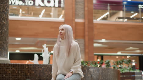 serene woman sits near indoor fountain in shopping mall scanning surroundings with gentle smile as ambient shoppers pass under bright lights near planters and benches for relaxing moments