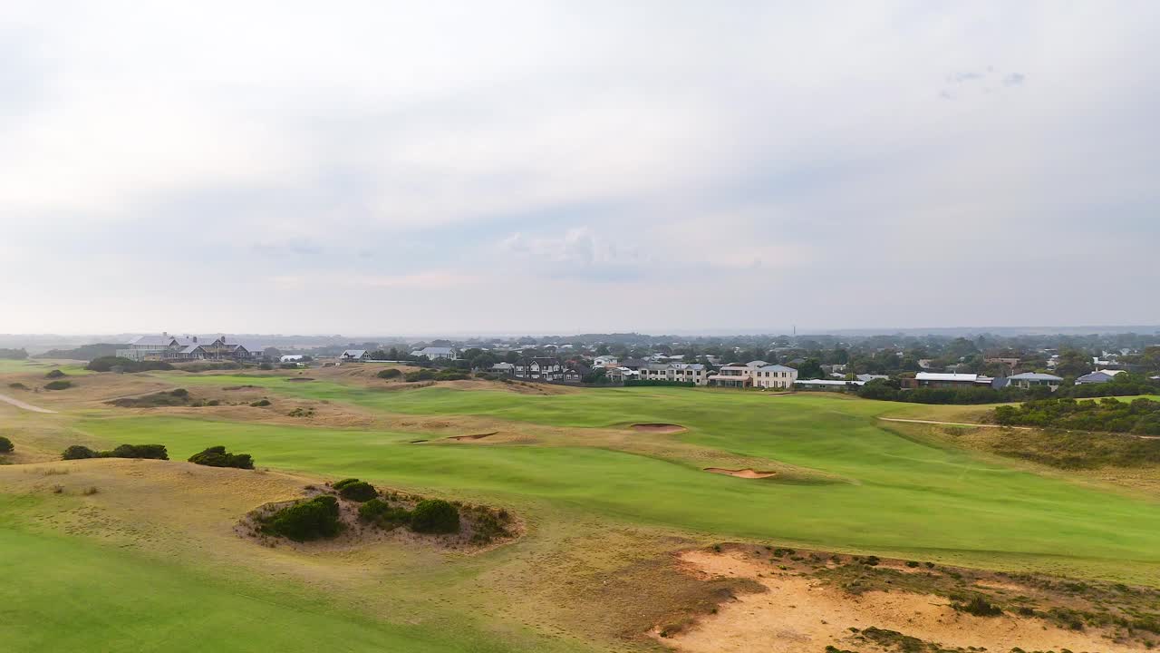 Aerial footage of Barwon Heads golf course with lush greens and sand traps under cloudy skies, showcasing expansive views