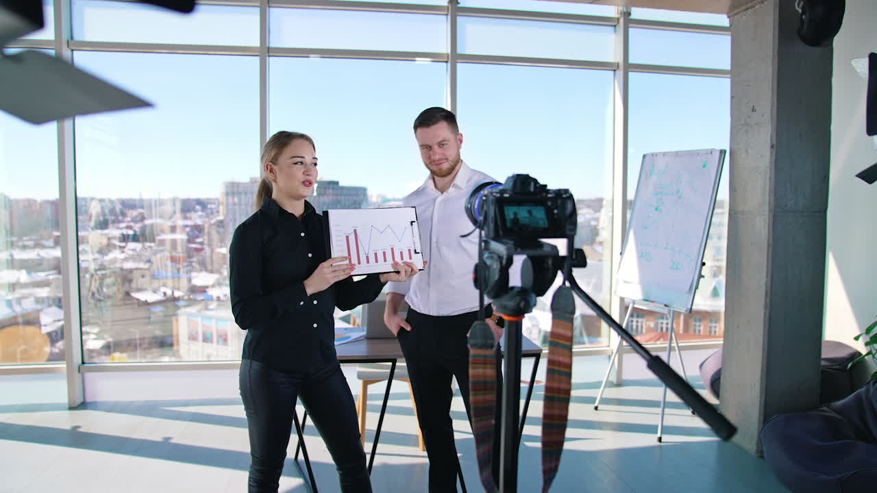 Attractive girl next to a young businessman records video. Young woman holding paper with graphs and speaking in front of professional camera in office room.