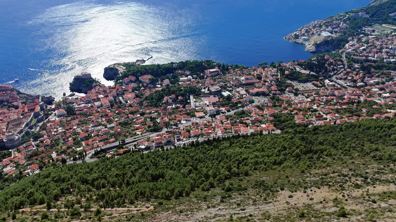 Aerial wide-angle drone faces outward over Dubrovnik’s Adriatic coastline, capturing terracotta rooftops, rocky shores, and the deep blue sea sparkling under bright sunlight along the coastline