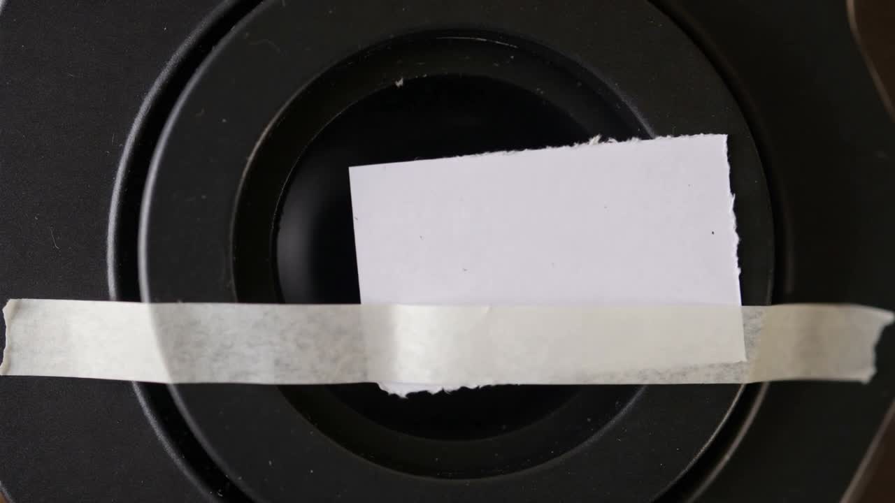 Shifting torn white paper square moving off black speaker cone on tabletop in room, tape loosening