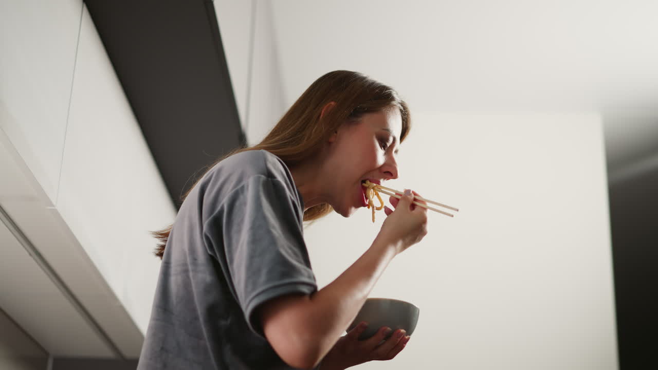 Side view of hungry lady eating dinner while standing in modern kitchen, using chopsticks to pick noodles from bowl, focused expression, soft bright background