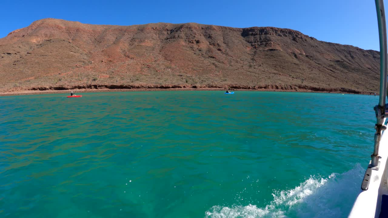 A moto boat cruises by a group of tourist kayakers paddling in teal blue water at the foot of a tropical mountain