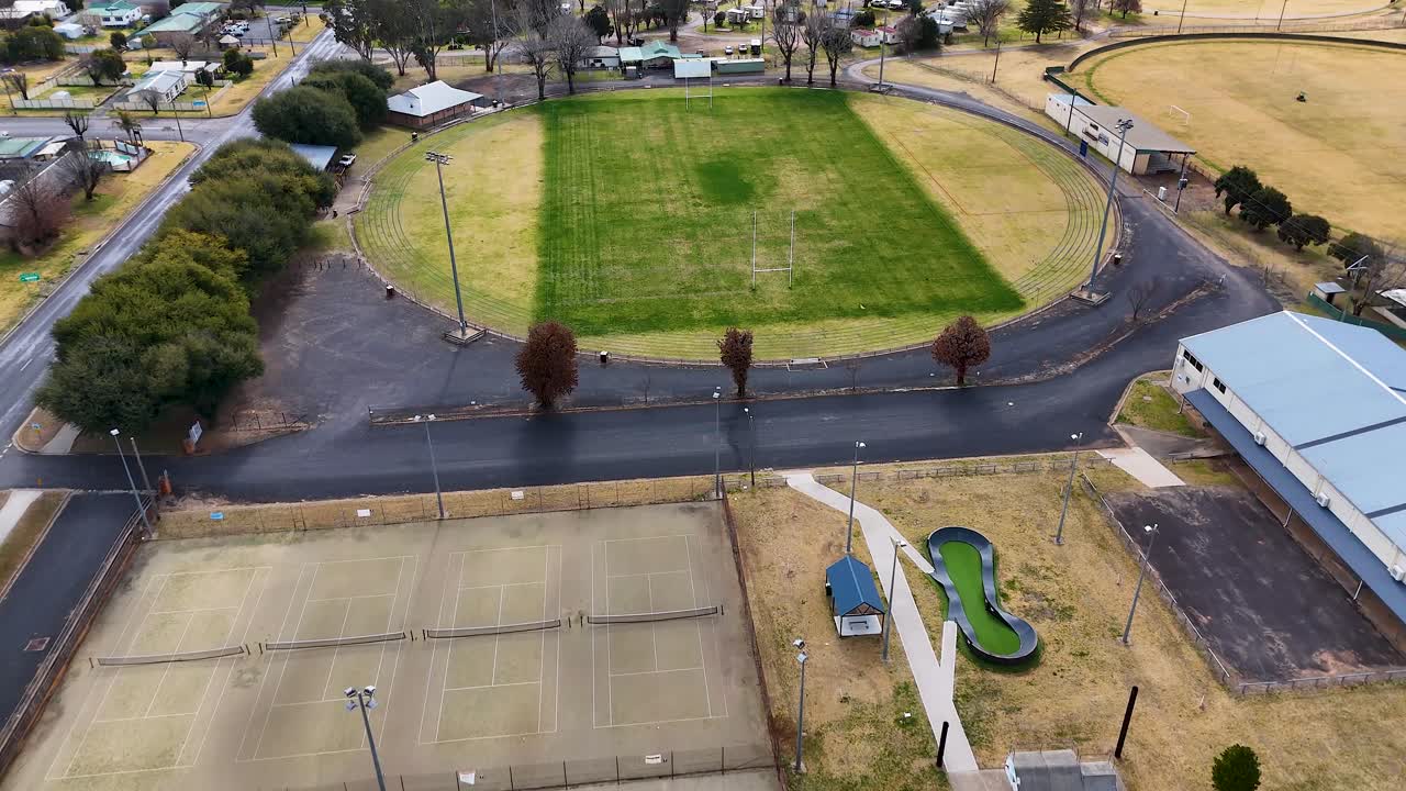 Aerial footage captures a gradual drone flyover above a green sports field, surrounded by residential homes, playground, and tennis courts in natural daylight