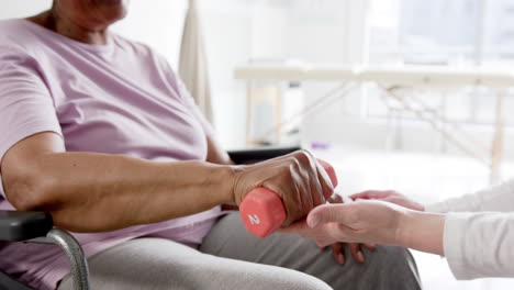 Diverse-senior-woman-exercising-with-weight-and-female-doctor-advising-in-hospital,-slow-motion