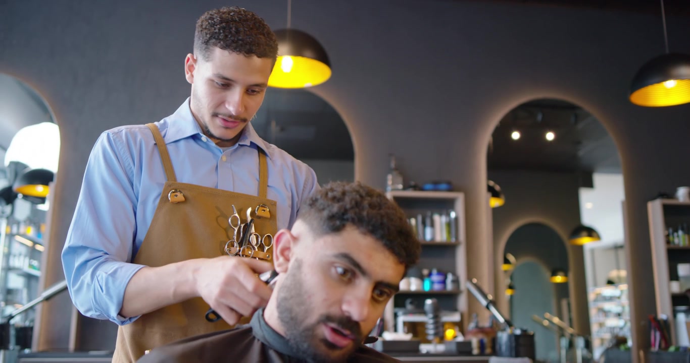Afroamerikansk mandlig frisør med forklæde klipper kunde efter hovedvip i salon, forfiner linje med trimmer.