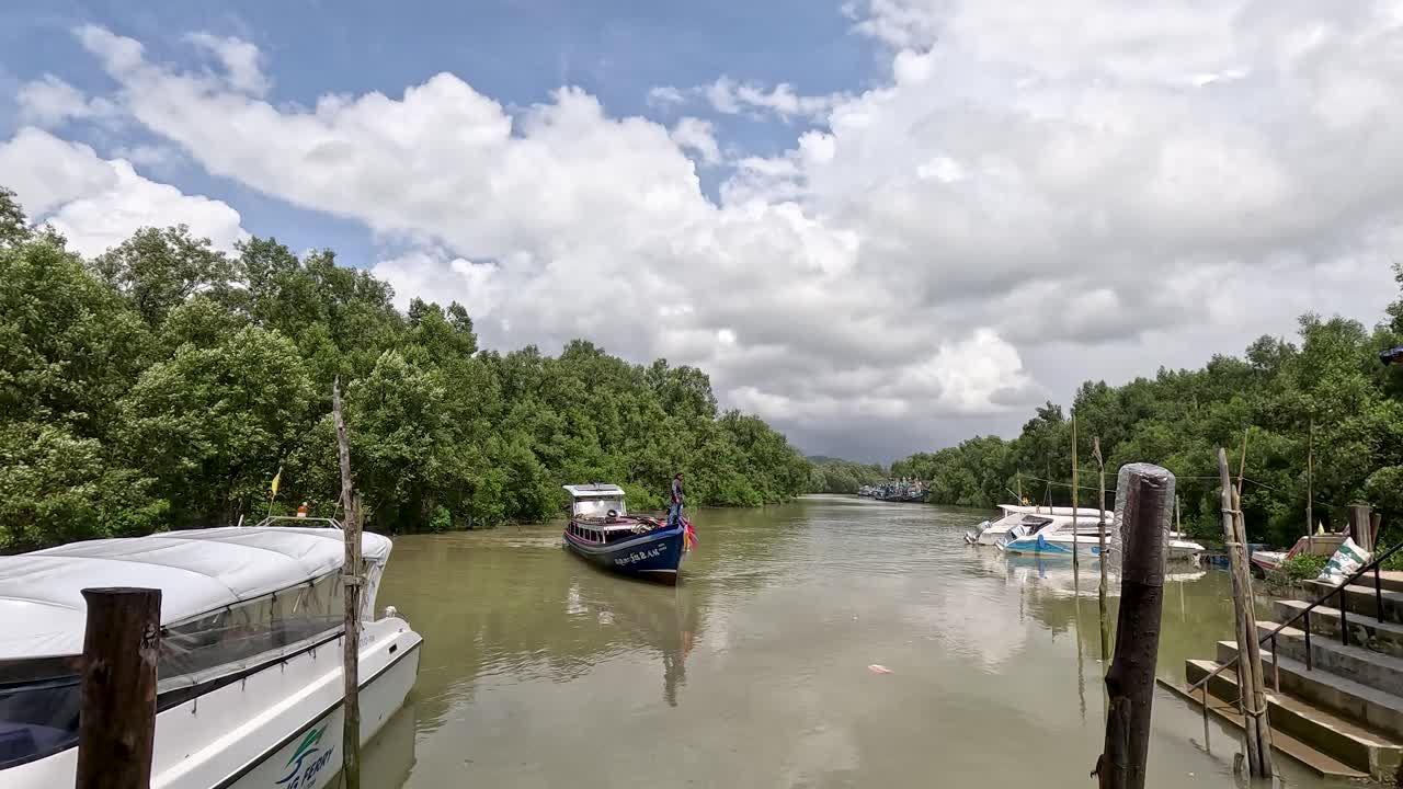 A speedboat travels along a calm mangrove river toward a wooden pier lined with boats under bright daylight, captured in a wide, steady shot