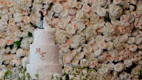 tiered wedding cake with couple topper in front of rose flower wall