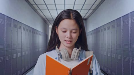 close up of asian teen girl student with a backpack holding and reading a book while standing in corridor