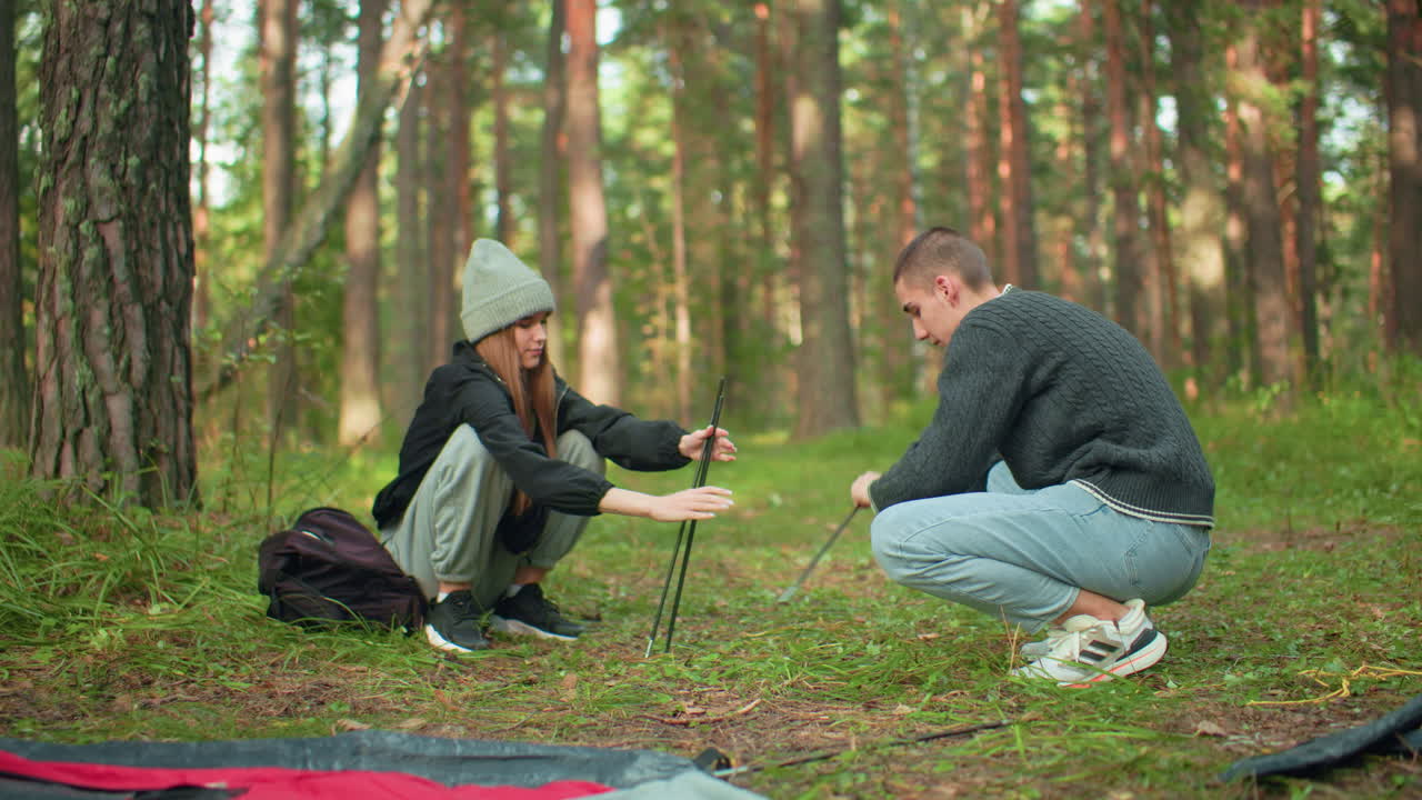 Couples squatting preparing tent for forest camping as lady hands pole to her love surrounded by camping materials, dry leaves, backpacks, and gear scattered around peaceful outdoor environment