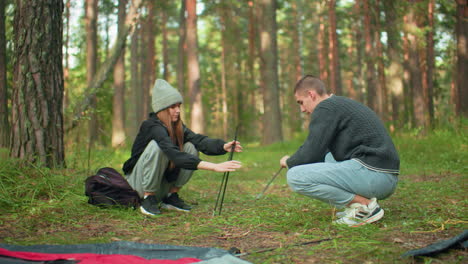 couples squatting preparing tent for forest camping as lady hands pole to her love surrounded by camping materials, dry leaves, backpacks, and gear scattered around peaceful outdoor environment