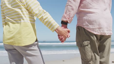 Mid-section-of-senior-caucasian-couple-holding-hands-on-the-beach