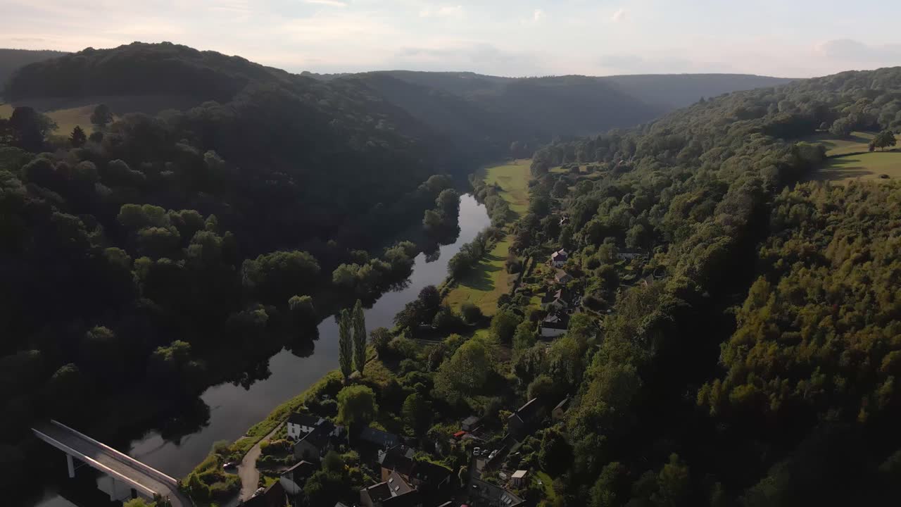 Aerial drone footage looking down on the picturesque village of Brockweir on a summers day, capturing it's historic stone bridge over the River Wye and surrounding lush greenery